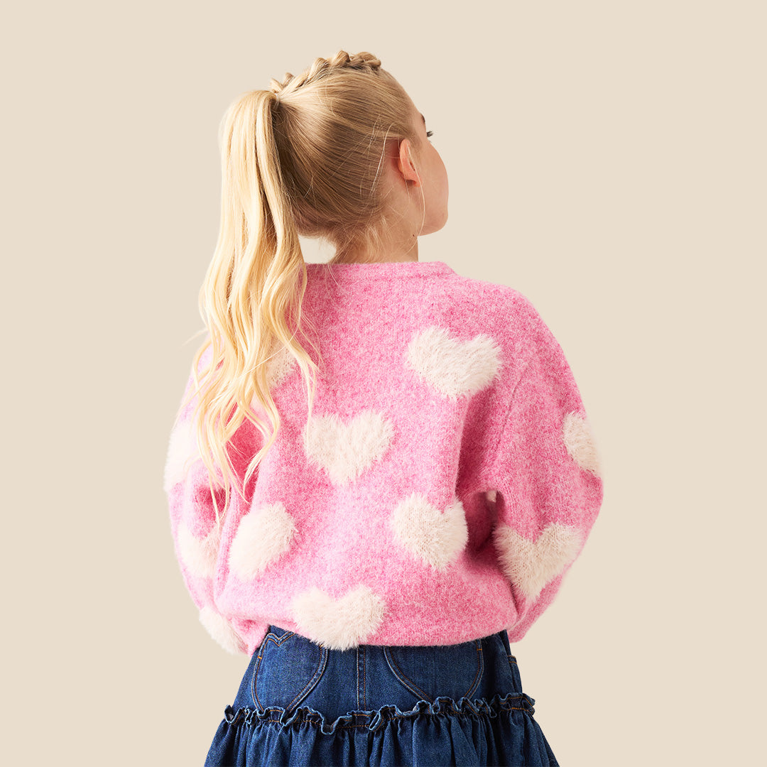 A young girl with blonde hair styled in buns wears a pink fuzzy knit sweater with fluffy white heart details, posing with her hands forming a heart shape in front of her chest. She is wearing a dark blue denim skirt.