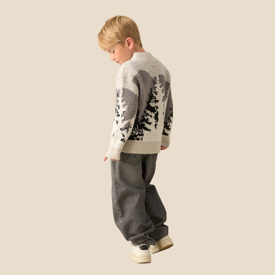 A young boy with blonde hair wearing a knit mock-neck sweater with a gray and black tree pattern, standing against a solid beige background.