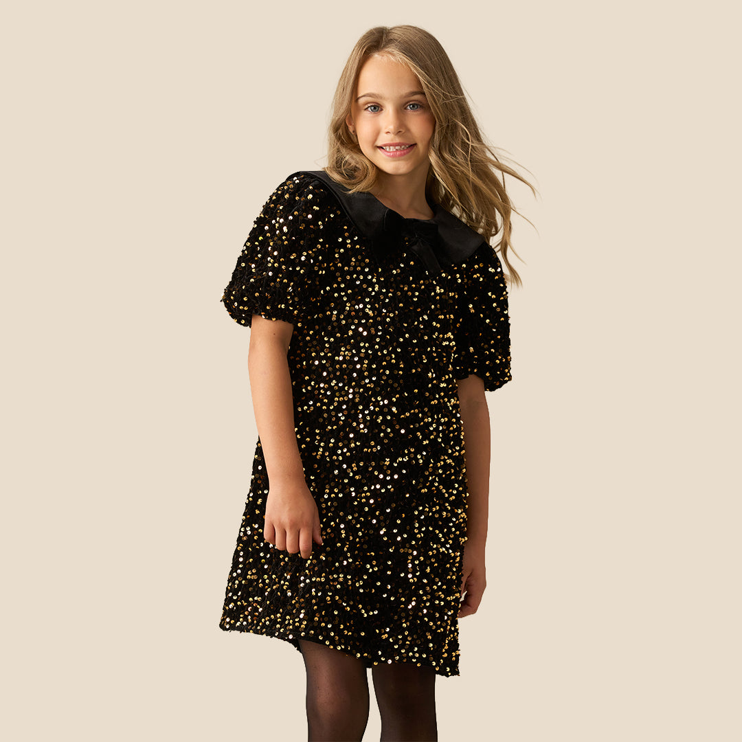 A young girl with blonde hair wearing a black short-sleeve dress with gold sequins and a black ruffled Peter Pan collar, standing against a solid beige background.
