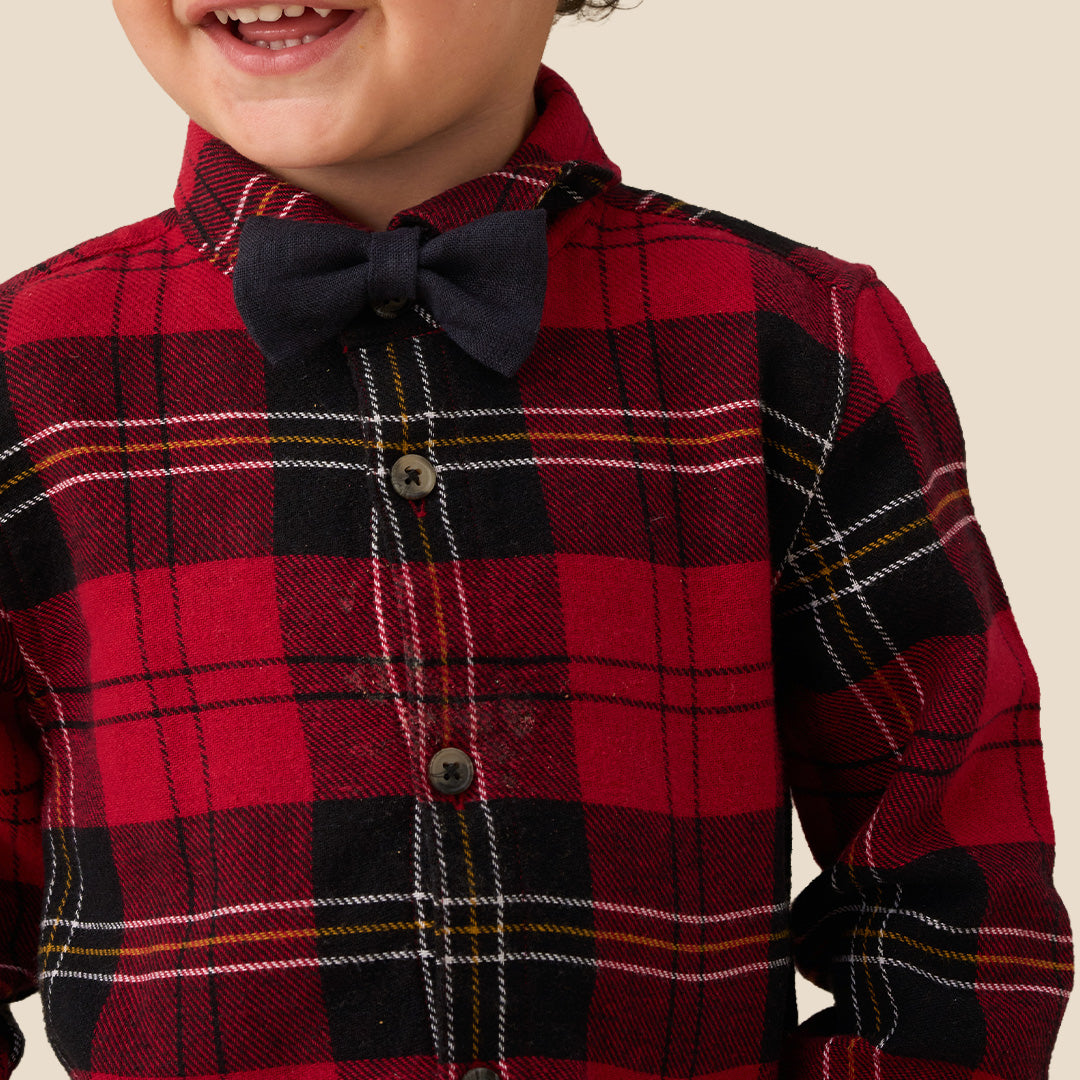 A toddler standing against a plain beige background, wearing a red and black plaid long-sleeve shirt with a matching dark bow tie, dark pants, and white sneakers.