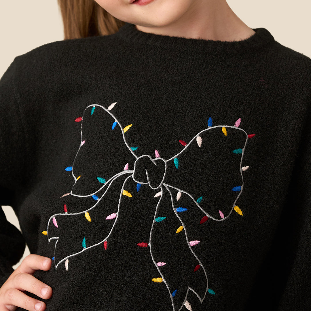 A young girl with her hair in a ponytail is wearing a black knit sweater featuring a large bow design outlined with colorful Christmas string lights, paired with a red pleated skirt, set against a solid beige background.