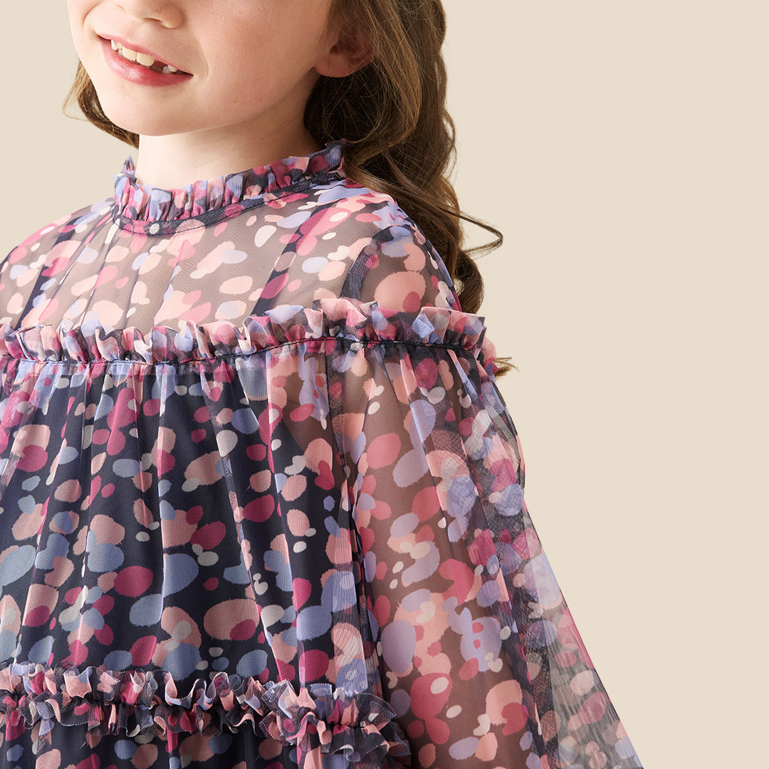 A young girl with curly hair stands on a checkered porch wearing a long-sleeved tiered dress with a pink, purple, and blue floral pattern. She is posing behind a black wrought-iron fence.