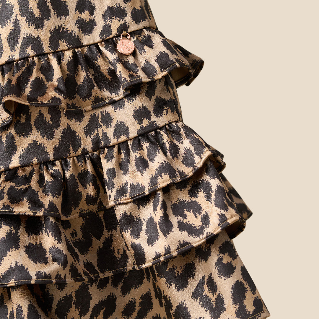 A young girl with blonde hair tied back wears a leopard print dress with puff sleeves and a tiered ruffle hem, standing against a solid beige background.