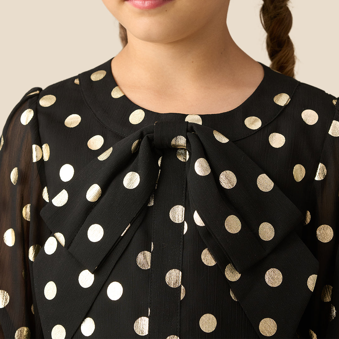 A young girl with braided hair wearing a black long-sleeve blouse with gold polka dots and black faux-leather pants, standing against a solid beige background.
