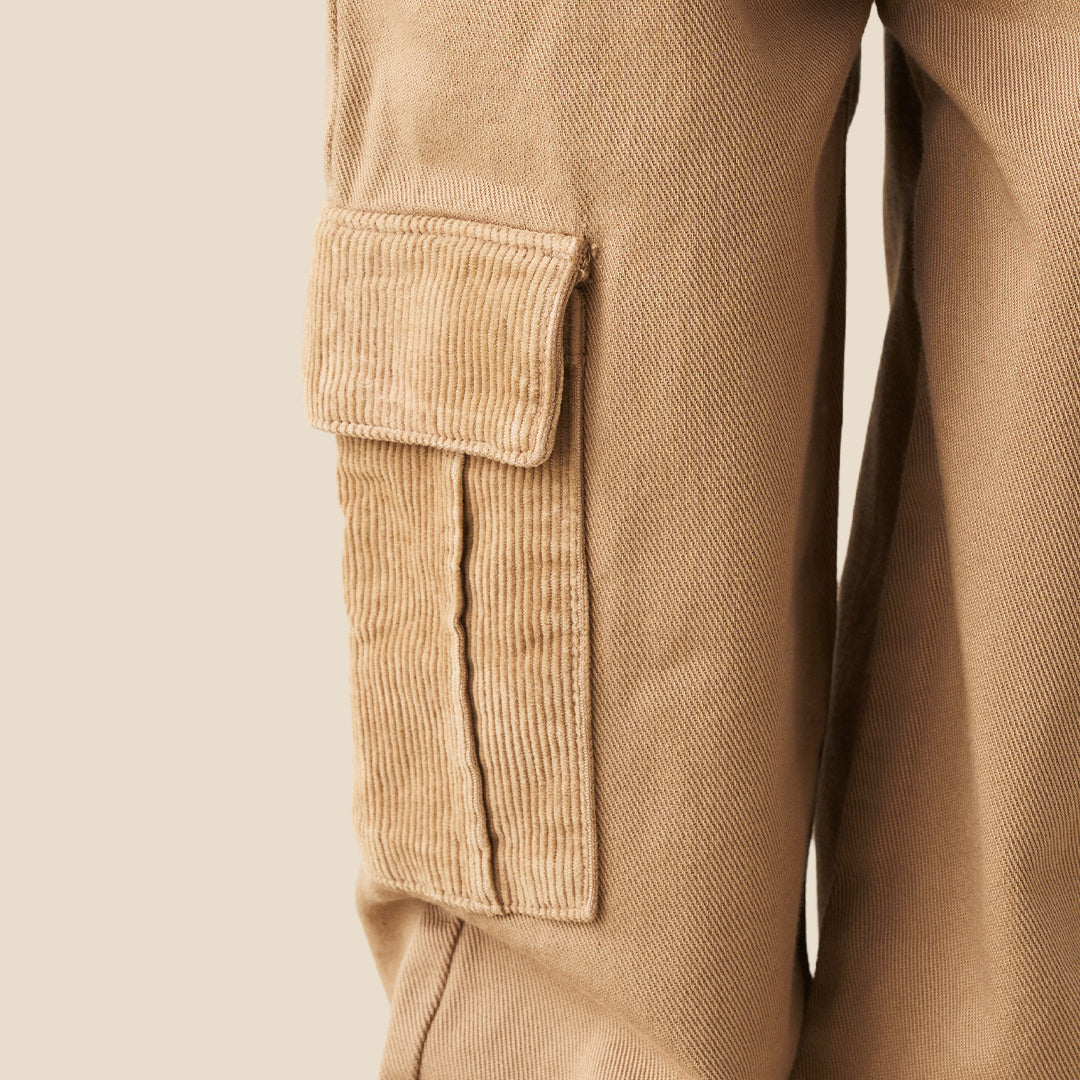 A child wearing wide-leg tan cargo pants with side flap pockets, styled with a light blue and white striped shirt and white sneakers against a solid beige background.