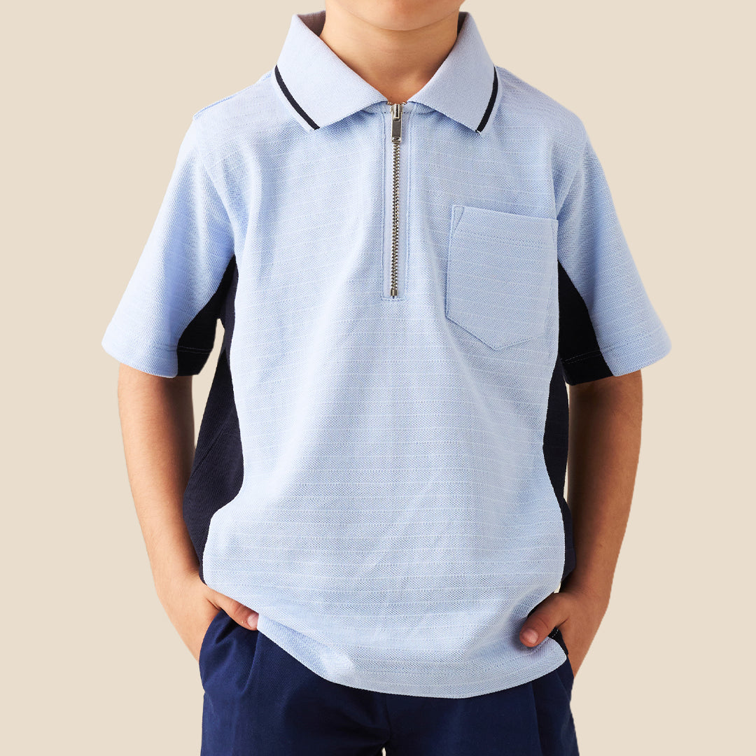 A young boy modeling a light blue polo shirt with dark navy side panels and a half-zip collar, paired with navy pants, against a solid beige background.