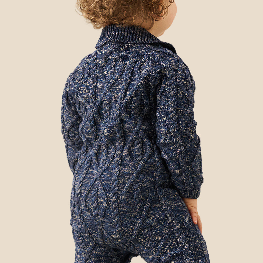 A baby with curly light brown hair wearing a navy blue cable-knit one-piece jumpsuit and white shoes, standing against a solid beige background.