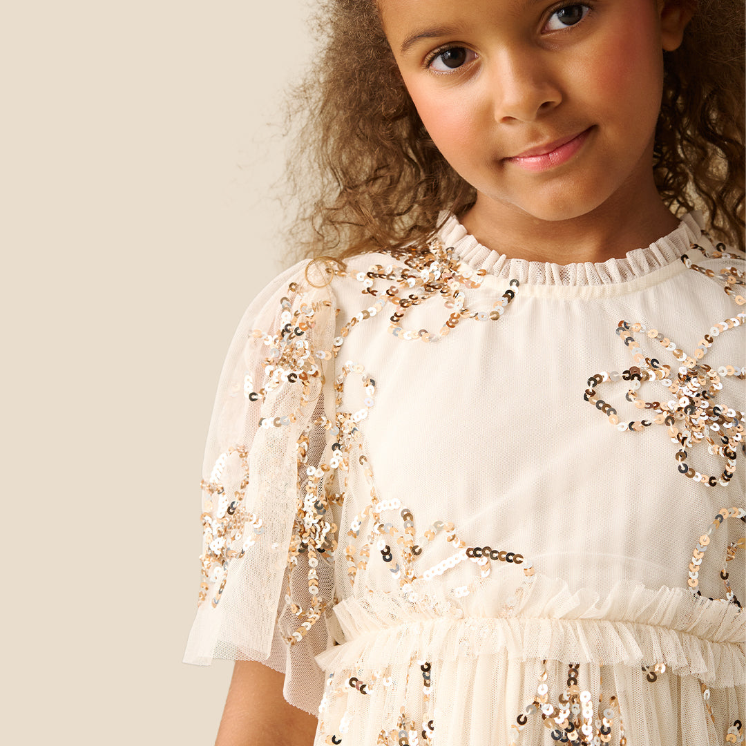 A young girl with curly brown hair wearing a tiered cream dress decorated with gold sequins and floral embroidery, standing against a solid beige background.