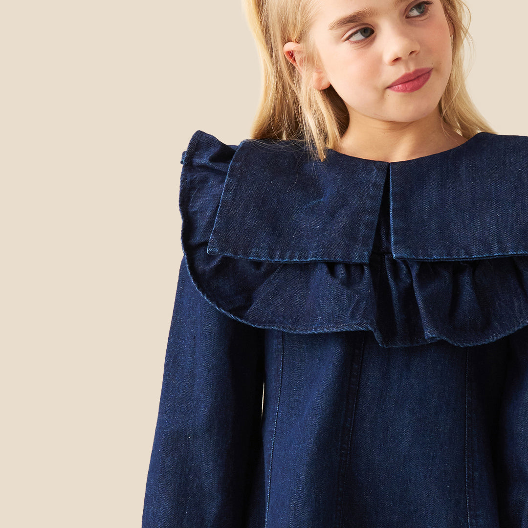 A young girl with blonde hair in two buns wearing a dark blue denim dress with a large ruffled collar, standing against a solid beige background.