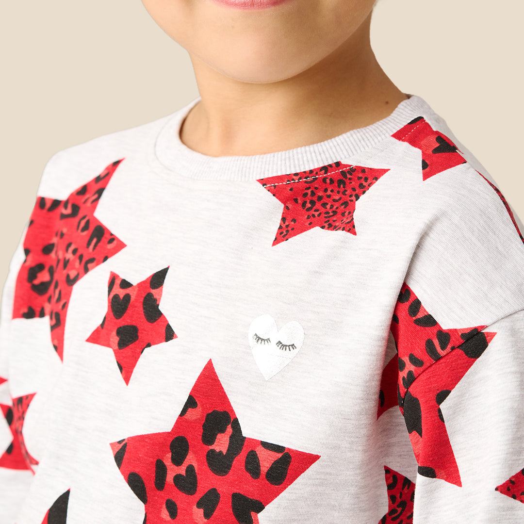 A young girl modeling a matching two-piece pajama set with a white base and a red star and ladybug print, set against a solid beige background.