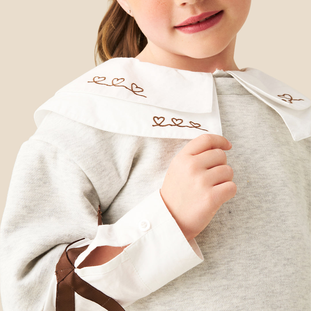 A young girl with her hair in a ponytail models a light gray long-sleeve top featuring a large white collar with brown embroidered details and matching brown accents on the sleeves, paired with blue jeans against a solid beige background.