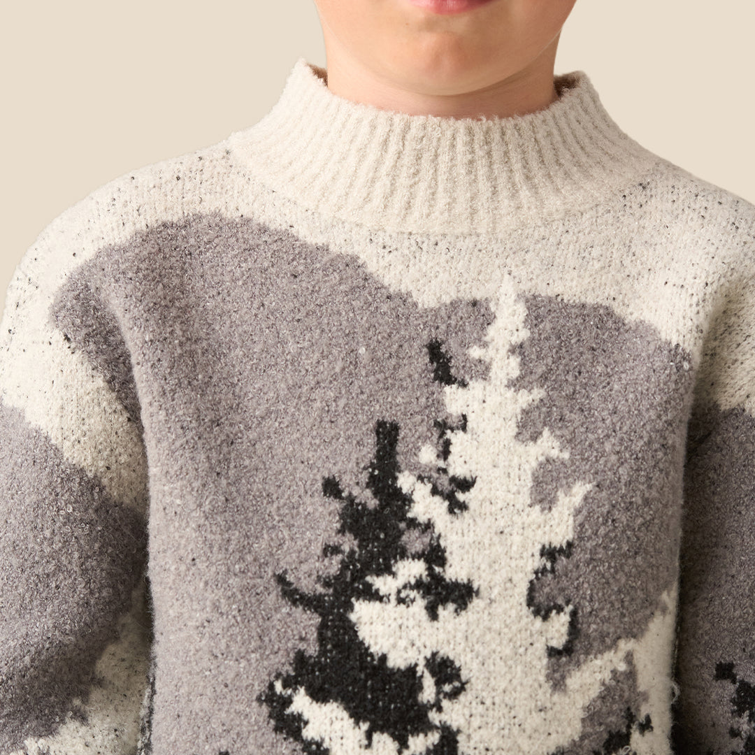 A young boy with blonde hair wearing a knit mock-neck sweater with a gray and black tree pattern, standing against a solid beige background.