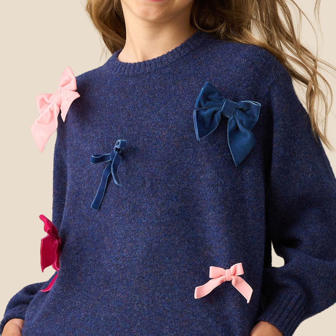 A young girl with blonde hair is smiling and posing in a navy blue knit sweater adorned with pink and blue fabric bows, paired with a red and black gingham pleated skirt, set against a solid beige background.