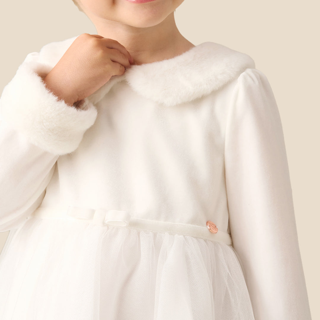 A young girl with blonde hair in a bun wearing a white long-sleeve tulle dress with faux fur trim on the collar and cuffs, standing against a solid beige background.