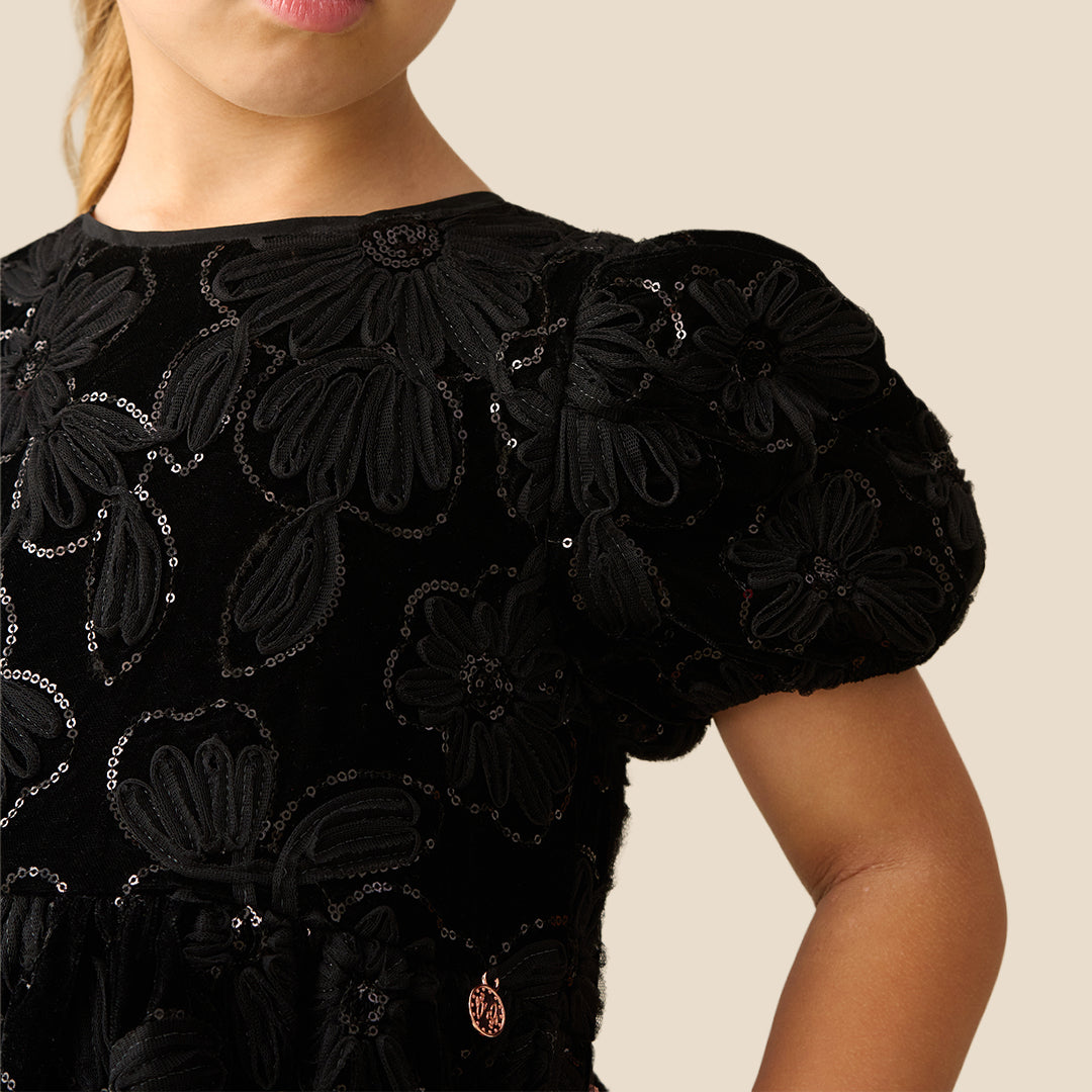 A young girl with her hair in a ponytail models a black short-sleeve dress with textured floral applique, standing against a solid beige background.