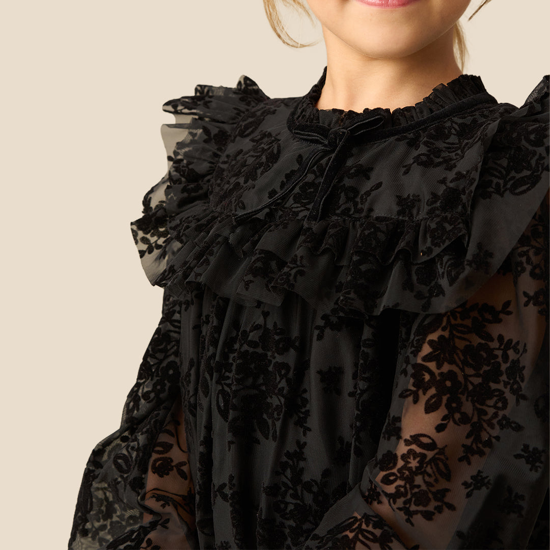 A young girl with blonde hair smiling while wearing a black lace long-sleeve dress with ruffled details, photographed against a solid light beige background.