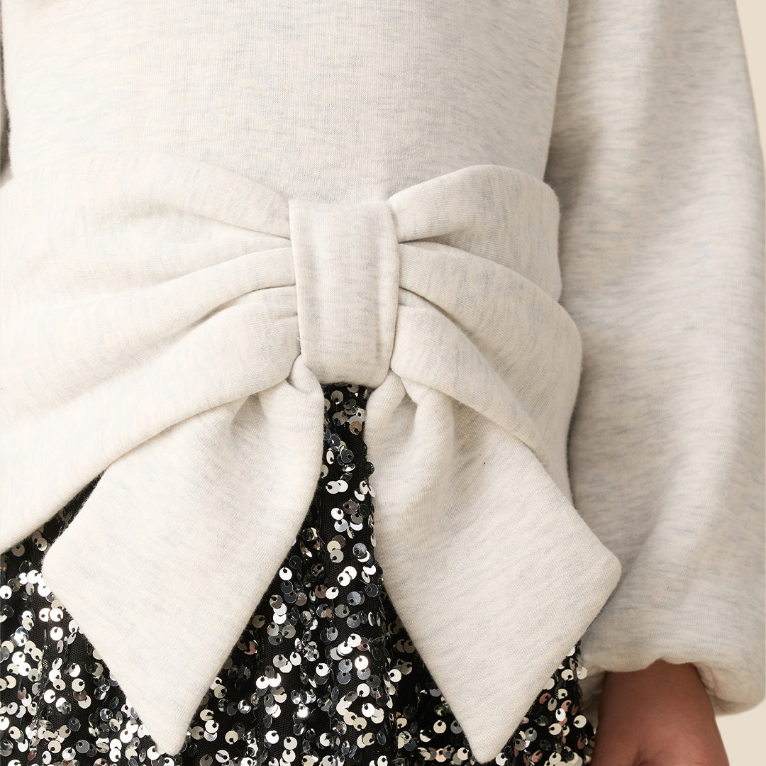 A young girl modeling a cream-colored knit sweater with a large matching fabric bow at the waist, paired with a black and white floral patterned skirt, against a solid beige background.