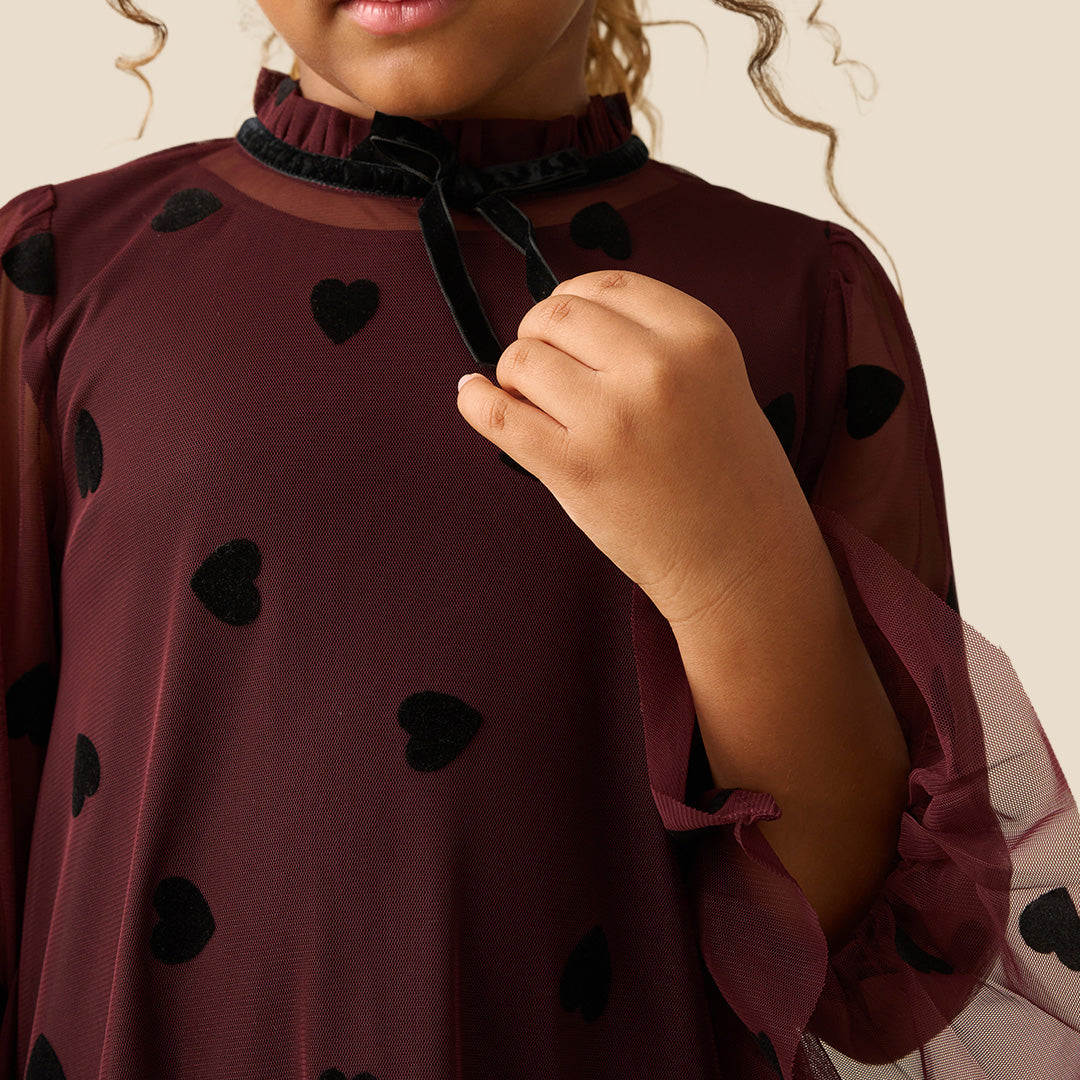 A young girl with curly brown hair wearing a burgundy sheer mesh dress with black heart patterns and a ruffled tulle hem, standing against a solid beige background.