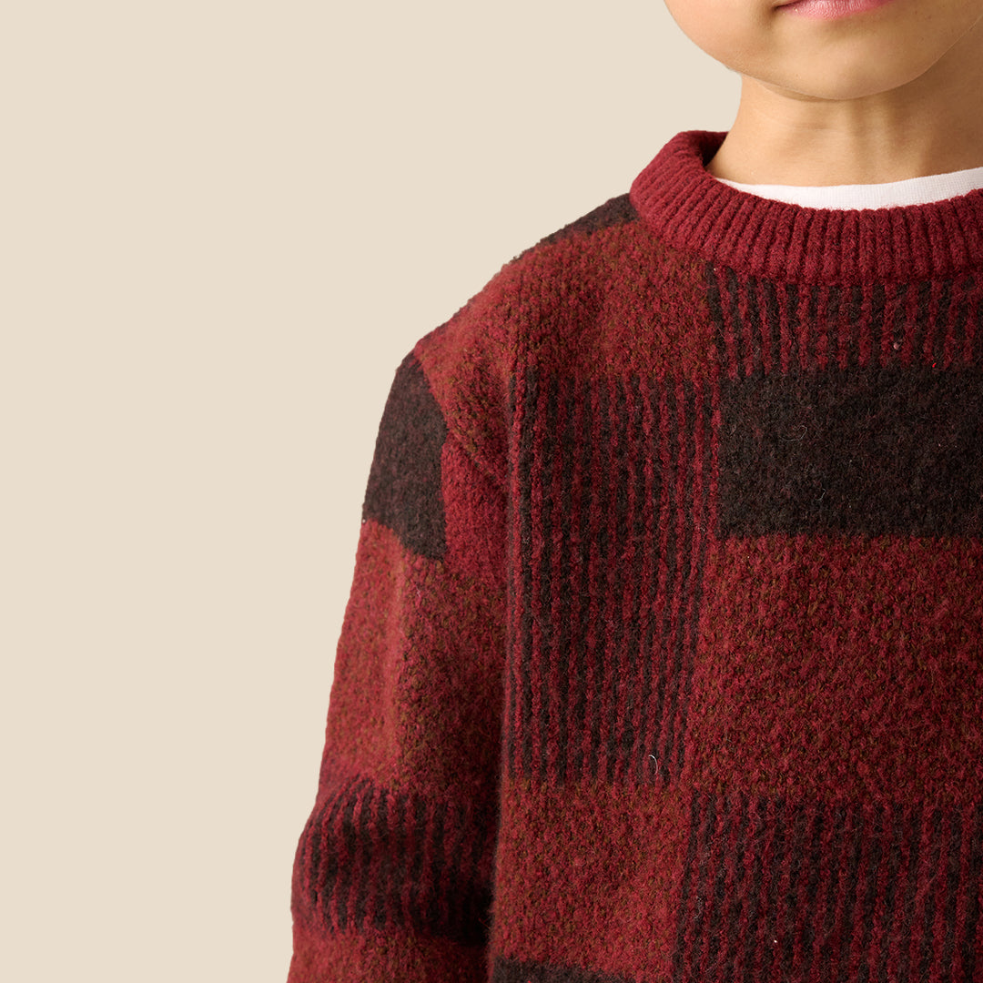 A young boy wearing a textured knit sweater with a dark red and black checkered pattern, paired with dark brown pants, set against a solid light beige background.