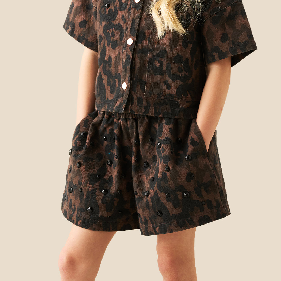 A child wearing a matching short-sleeve button-front top and shorts set in a dark brown and black leopard print, with decorative pearl-like embellishments, photographed against a plain beige background.