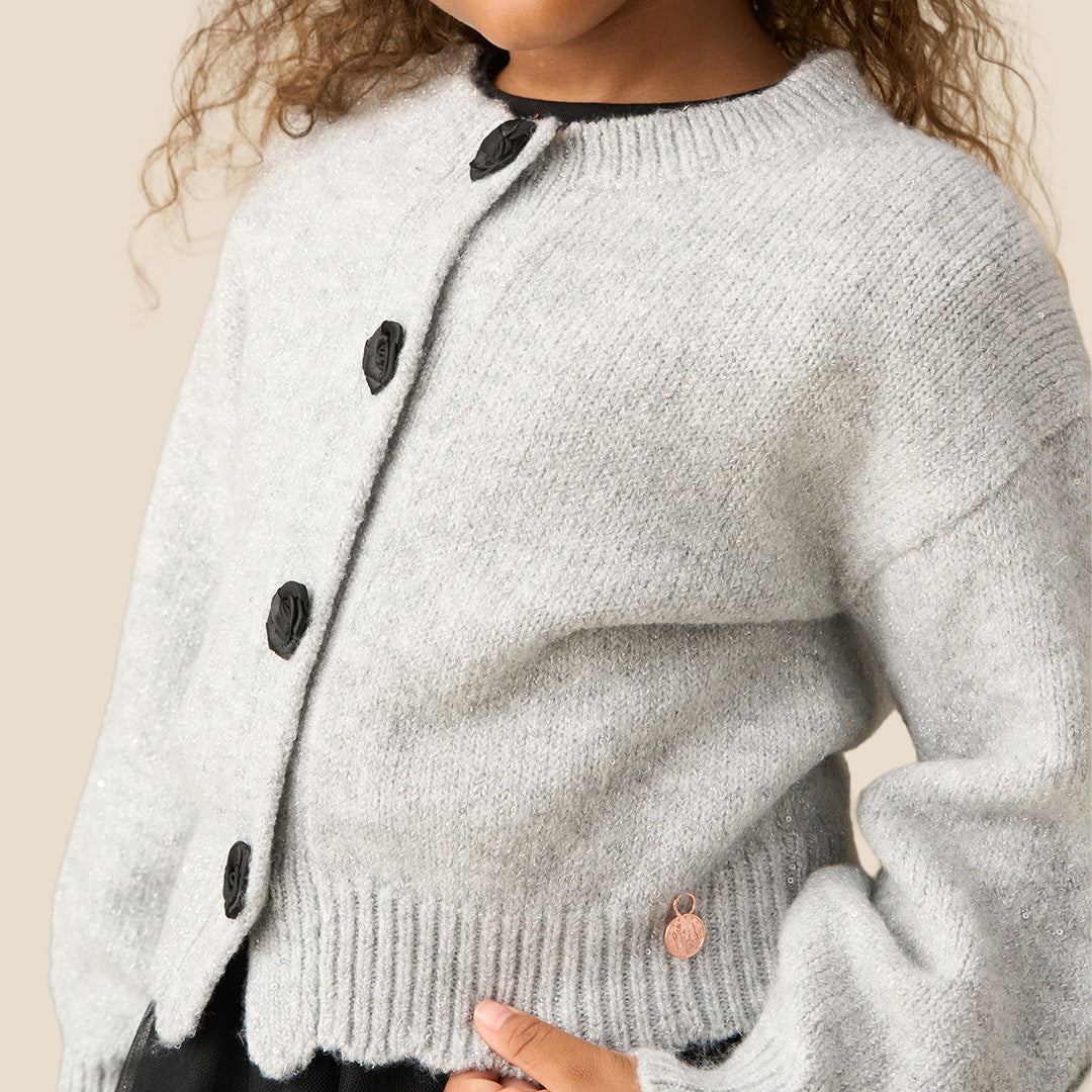 A young girl with curly brown hair wearing a light gray knit cardigan with black buttons and a black ruffled skirt, standing against a solid beige background.