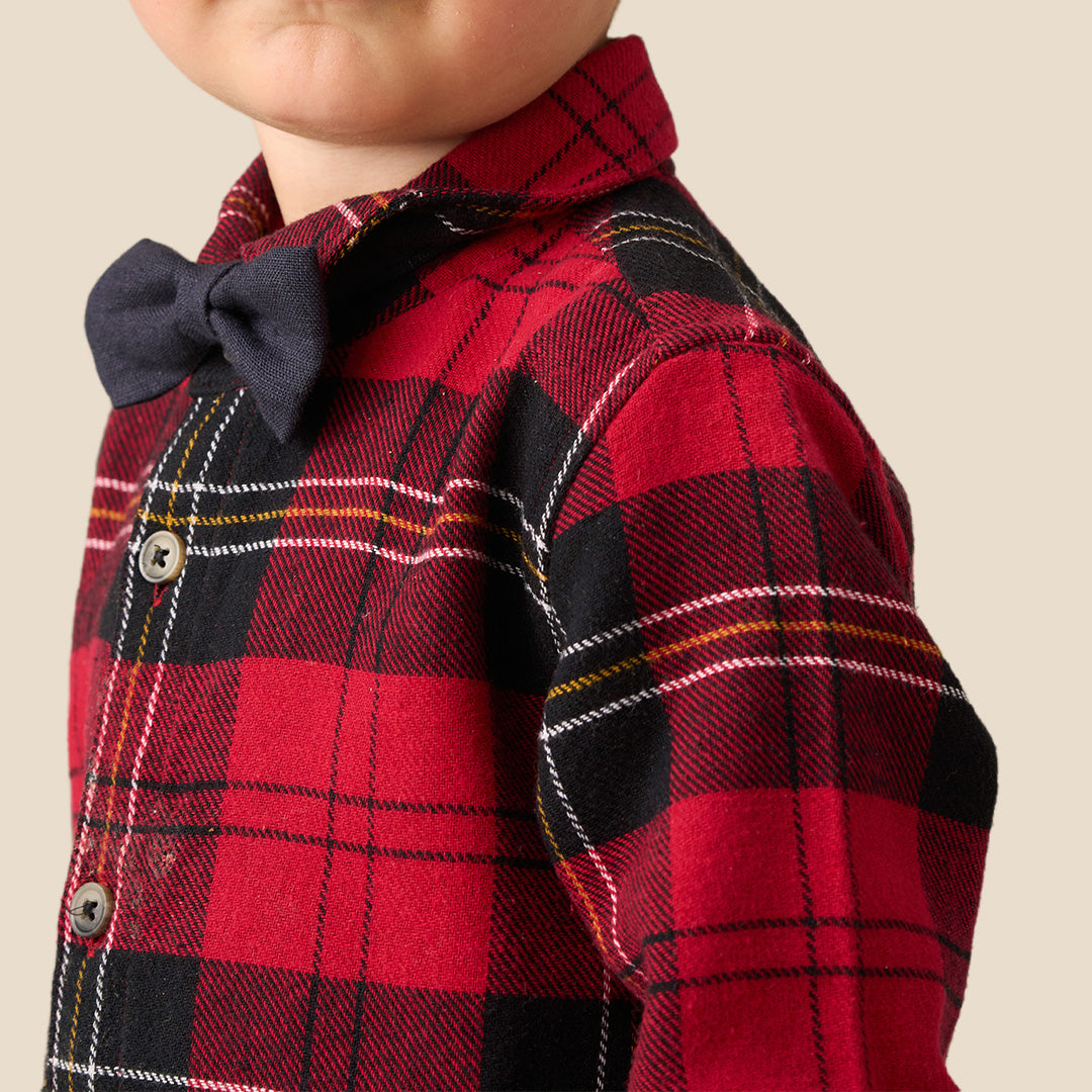 A toddler standing against a plain beige background, wearing a red and black plaid long-sleeve shirt with a matching dark bow tie, dark pants, and white sneakers.