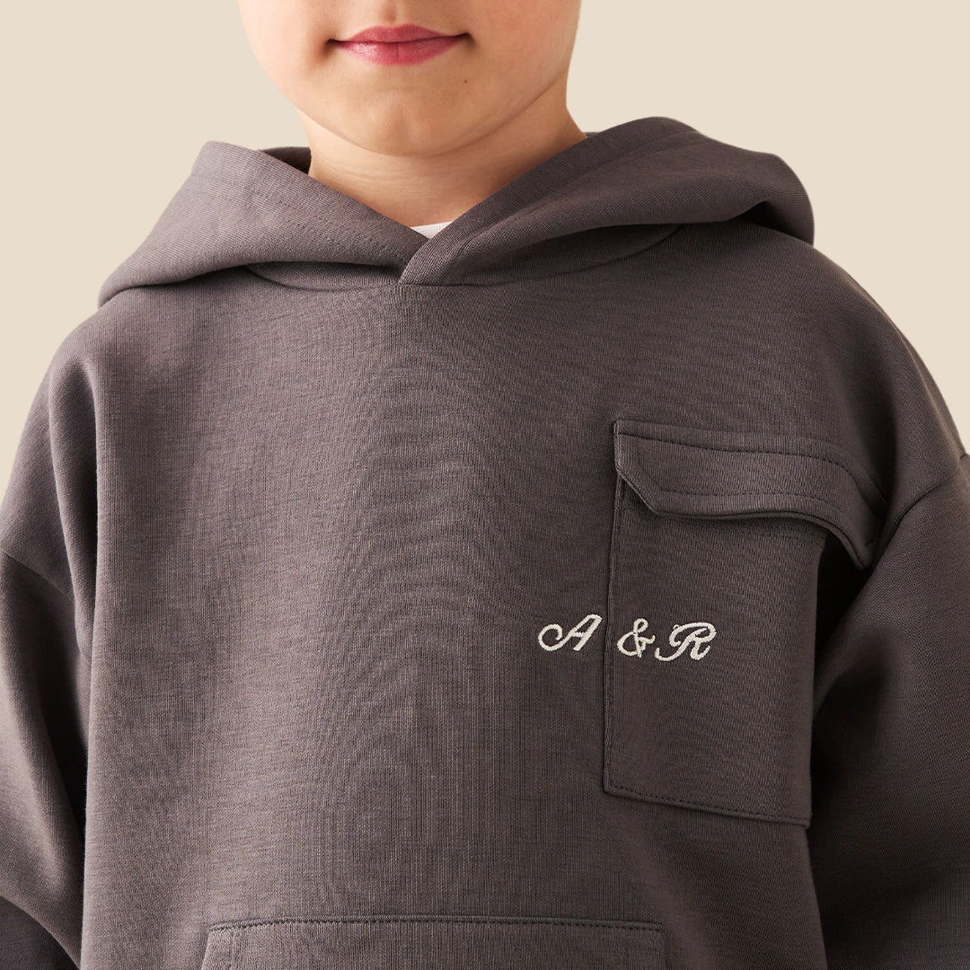 A young boy wearing a dark gray hoodie with a small white graphic on the chest and matching dark gray cargo pants, standing against a solid beige background.