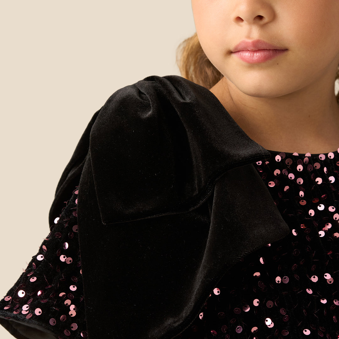 A young girl with light brown hair in pigtails stands against a solid beige background wearing a short-sleeved black dress covered in small pink and white floral patterns, with a black bow on the shoulder.