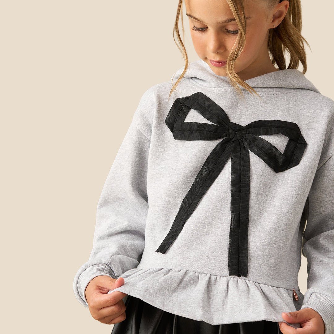 A young girl with her hair in a ponytail models a light gray heathered hoodie featuring a large black bow graphic on the front and a ruffled hem, paired with a black faux-leather skirt, against a solid beige background.