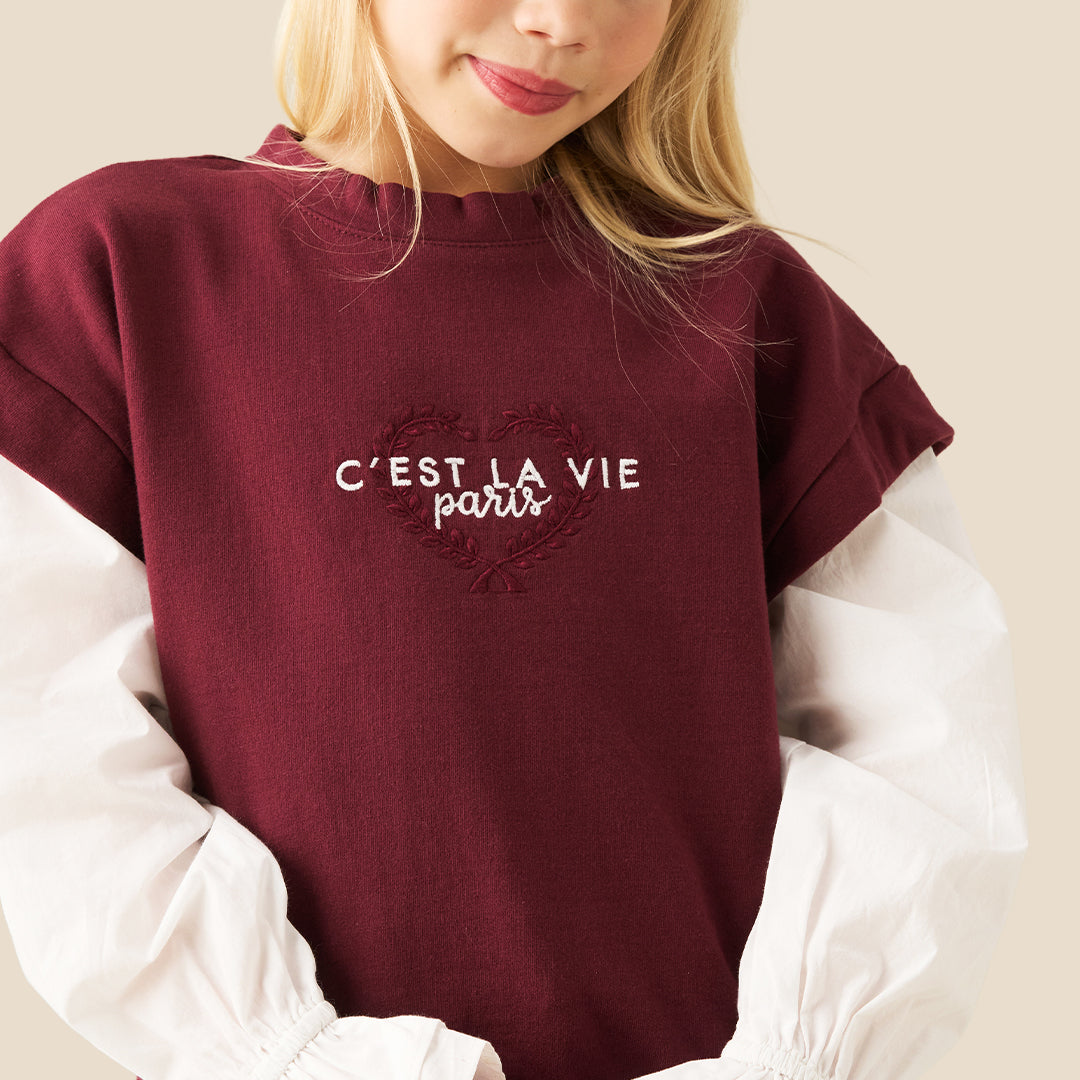 A young girl with blonde pigtails wearing a maroon cropped t-shirt with white sleeves and 'C'EST LA VIE paris' printed in white, layered over a white long-sleeve blouse with ruffled cuffs, and blue jeans, against a solid beige background.