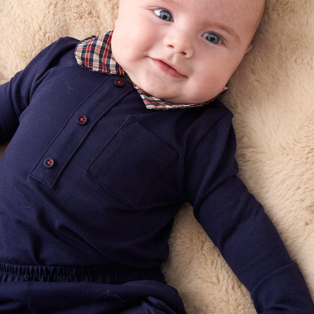 A smiling toddler with curly blond hair wearing a navy long-sleeve shirt and matching pants over a red, white, and blue plaid collared shirt, photographed against a plain warm beige studio background.