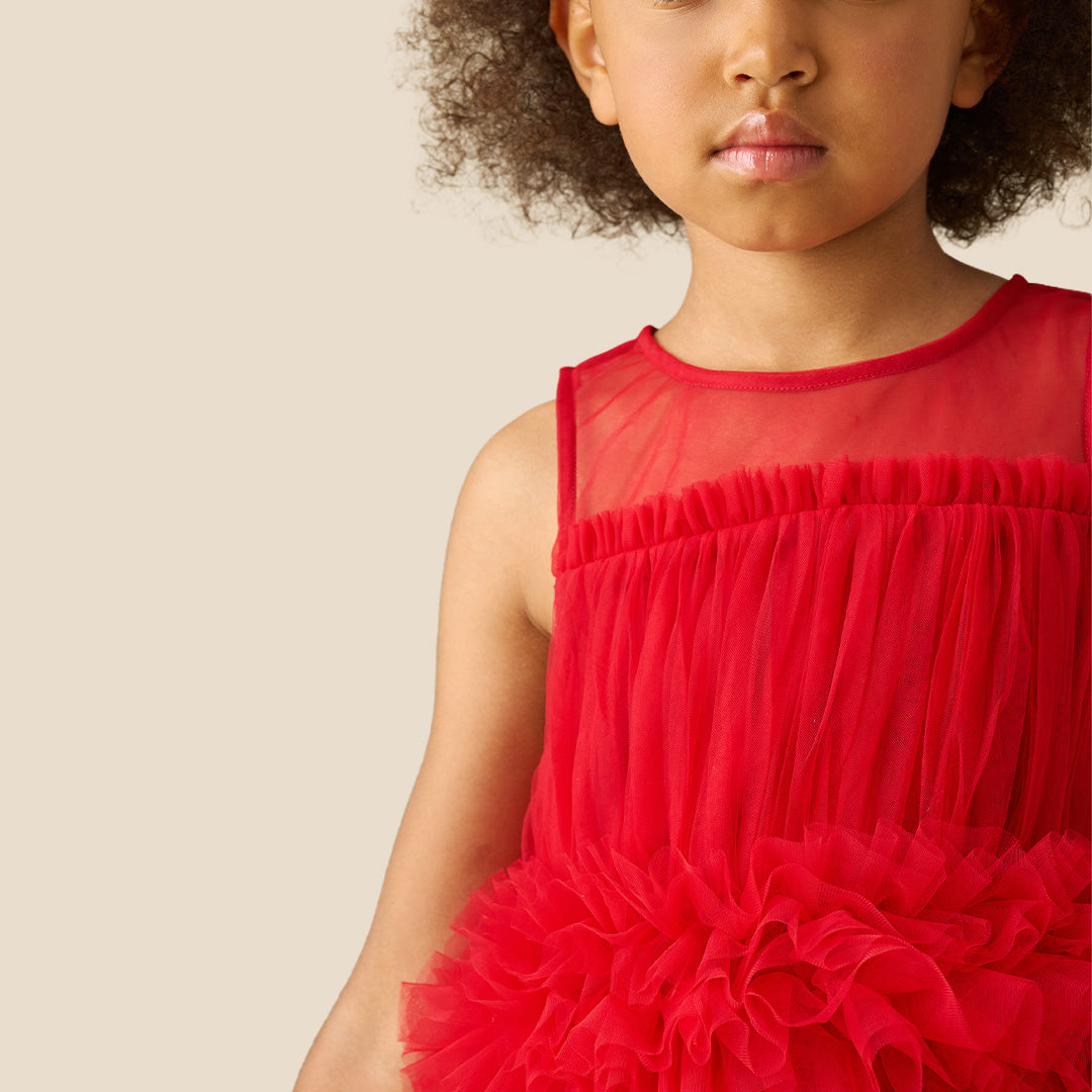 A young girl with curly brown hair wearing a sleeveless bright red tulle dress with a ruffled waistband, standing against a solid beige background.