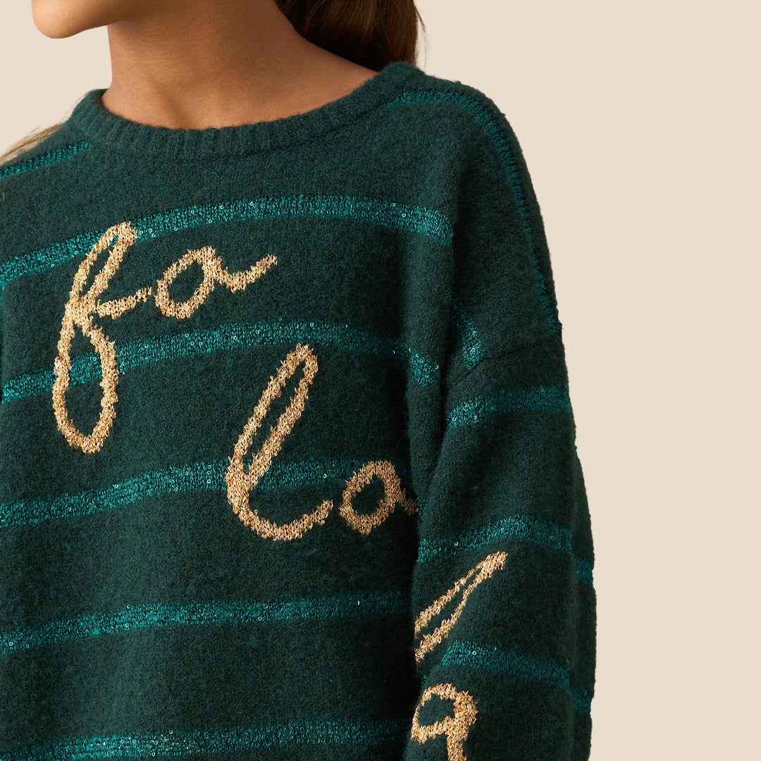 A young girl smiling while wearing a dark green knit sweater with teal horizontal stripes and gold glittery 'fo'lo' lettering on the front, paired with a gold sequined skirt, against a solid beige background.