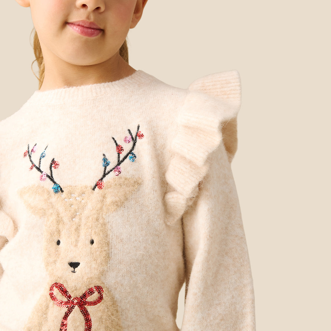 A young girl with blonde hair in a ponytail models a cream-colored knit sweater featuring a large embroidered reindeer on the front with colorful antlers and a red bow. She is wearing a shiny red skirt against a solid light beige background.