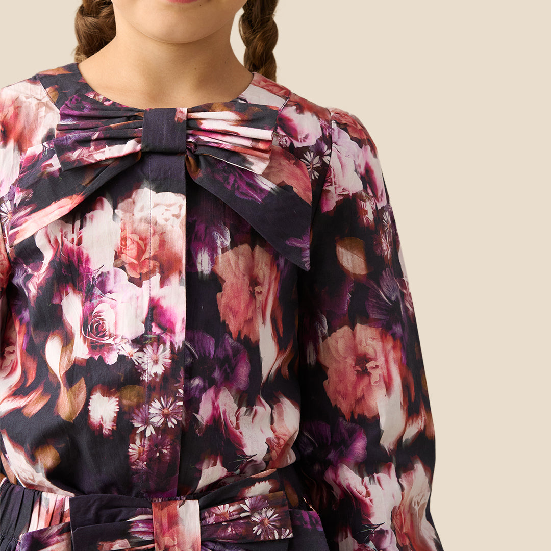 A young girl with braided hair wearing a long-sleeve floral blouse with a large bow at the collar, photographed against a plain light beige studio background.