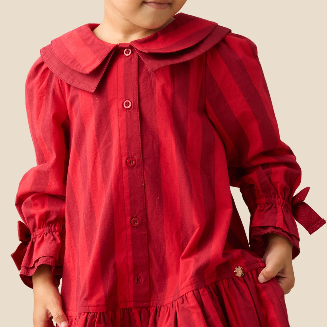 A child wearing a red long-sleeve dress with vertical stripes, a large collar, and tie details on the cuffs, shown against a plain beige background.