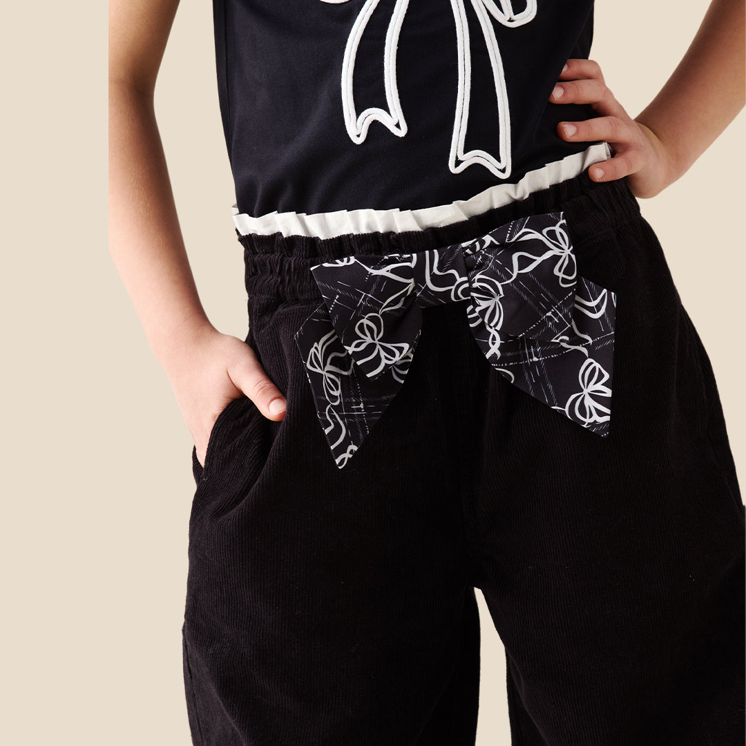 A child wearing black wide-leg pants with a white bow and floral pattern at the waistband, paired with a black top and red sneakers, standing against a solid beige background.