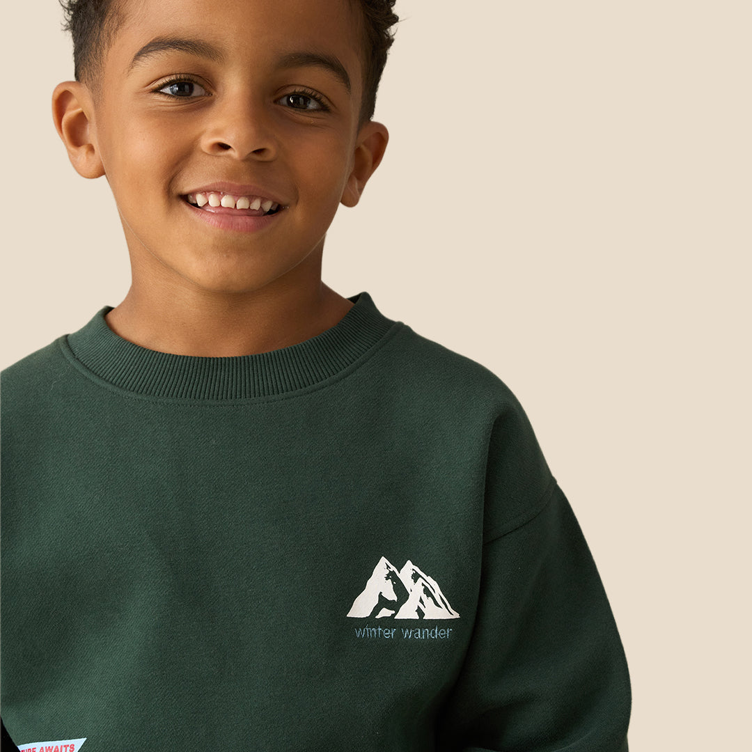 A young boy with curly dark hair wearing a dark green crewneck sweatshirt with a small white graphic on the left chest and khaki pants, posing against a solid light beige background.