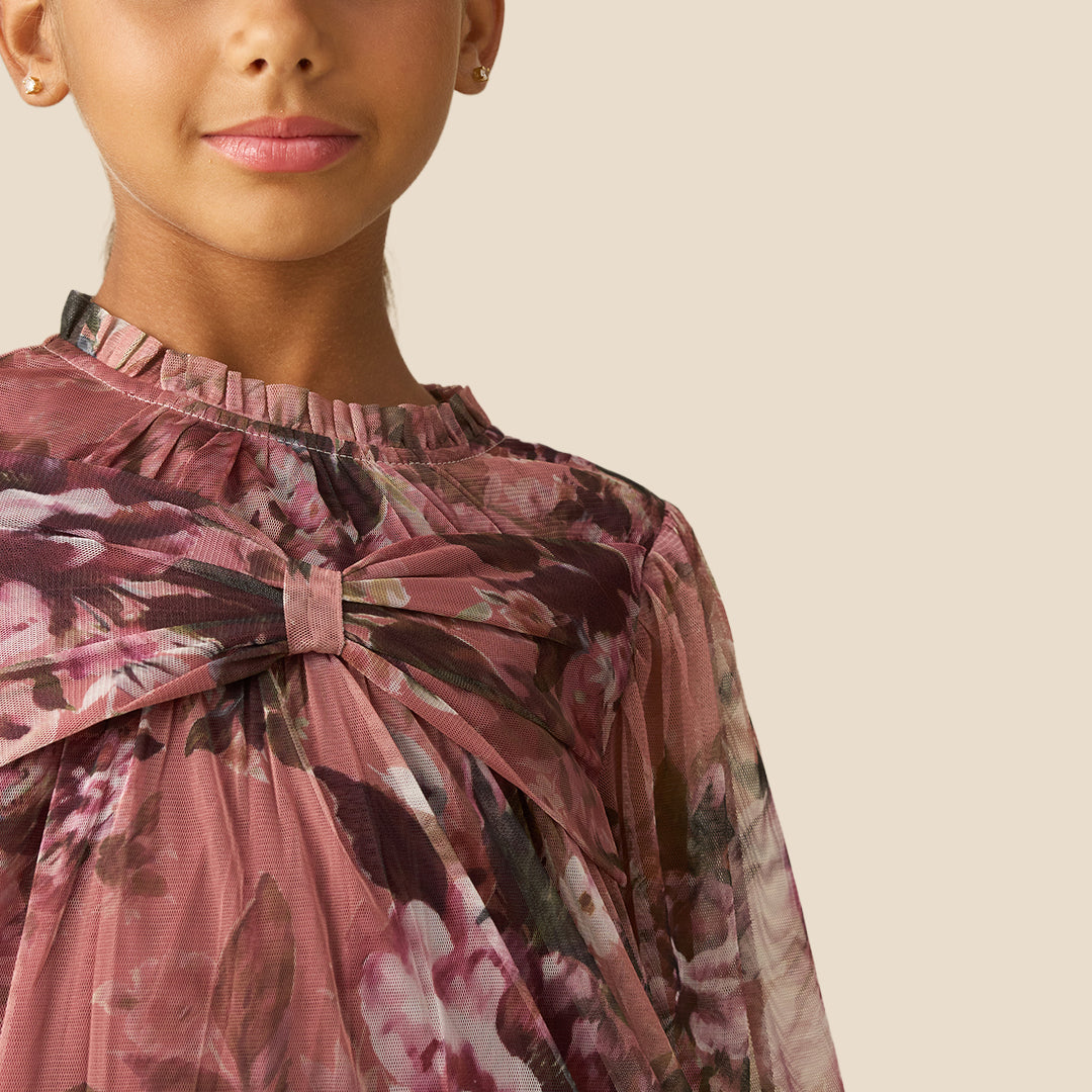 A young girl with her hair in a ponytail models a long-sleeve tiered dress with a dusty rose and brown floral print against a solid beige background.