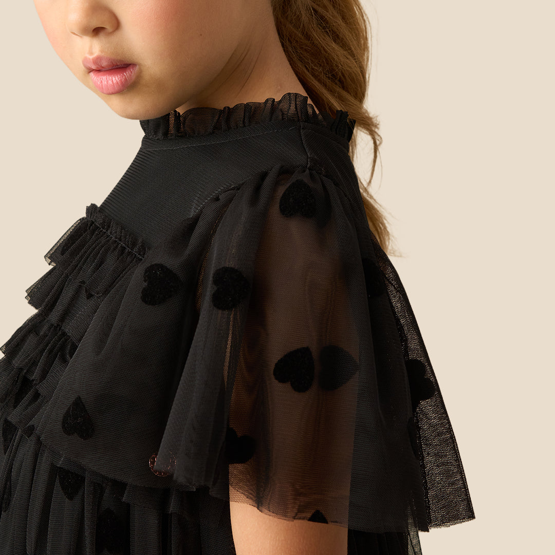 A young girl with her hair in a ponytail models a black tulle dress with heart patterns and sheer flutter sleeves, set against a solid beige background.