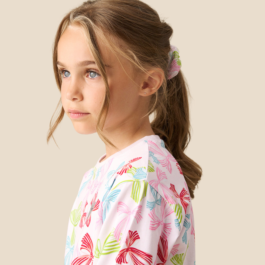 A young girl modeling a two-piece pajama set featuring a long-sleeve top and matching pants with a colorful bow pattern on a light pink background, set against a solid beige backdrop.