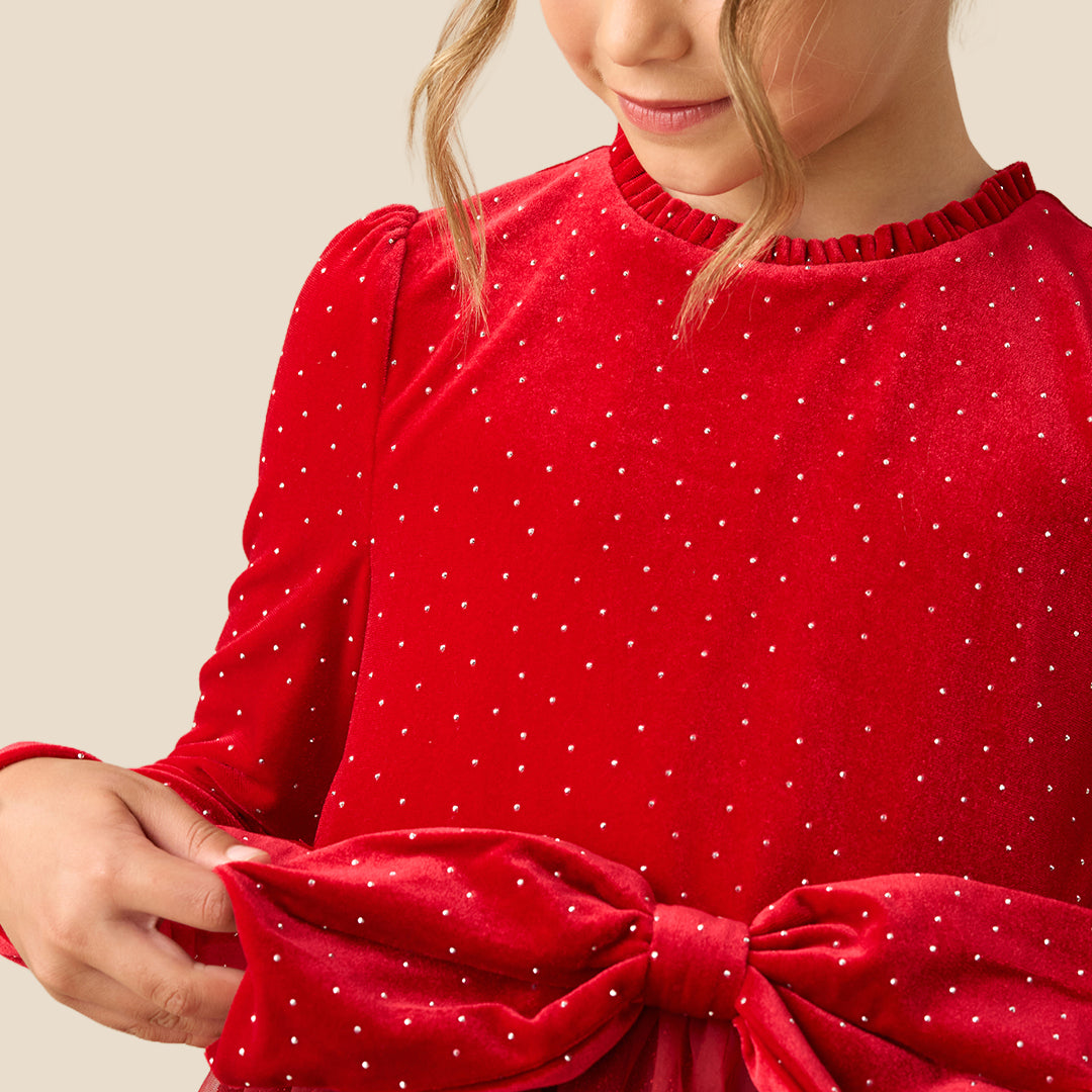 A young girl wearing a festive red dress with long sleeves, tiny gold polka dots, a matching red bow at the waist, and a tulle skirt, set against a solid beige background.