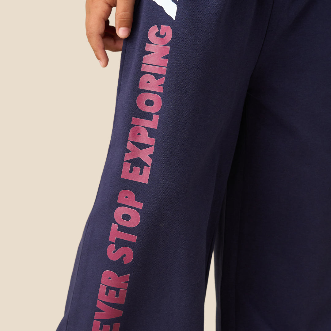 A young boy modeling a two-piece pajama set consisting of a bright blue long-sleeve shirt and navy blue wide-leg pants with pink text on the leg, set against a solid beige background.