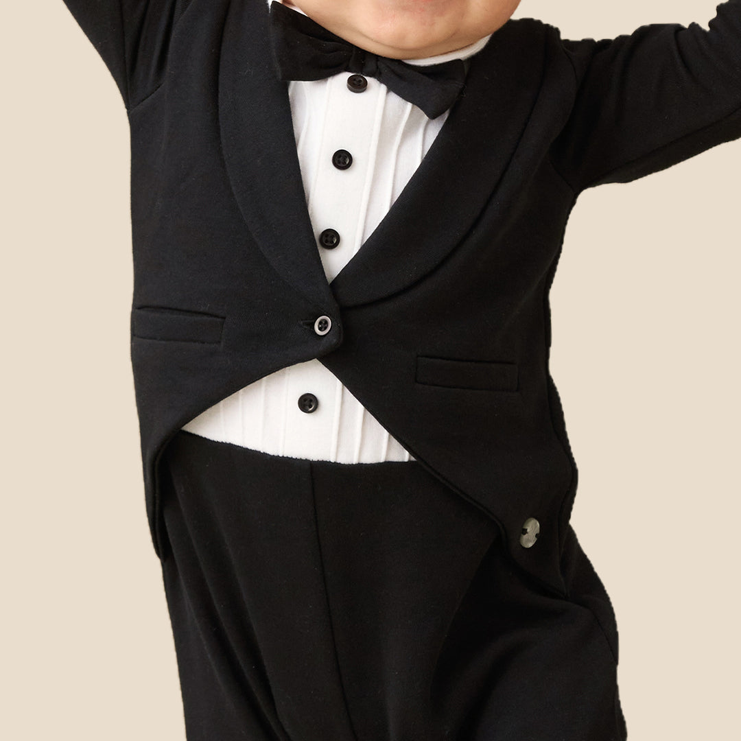 A baby wearing a black formal tuxedo-style romper with a white shirt and black bow tie, posing with arms raised against a plain beige background.