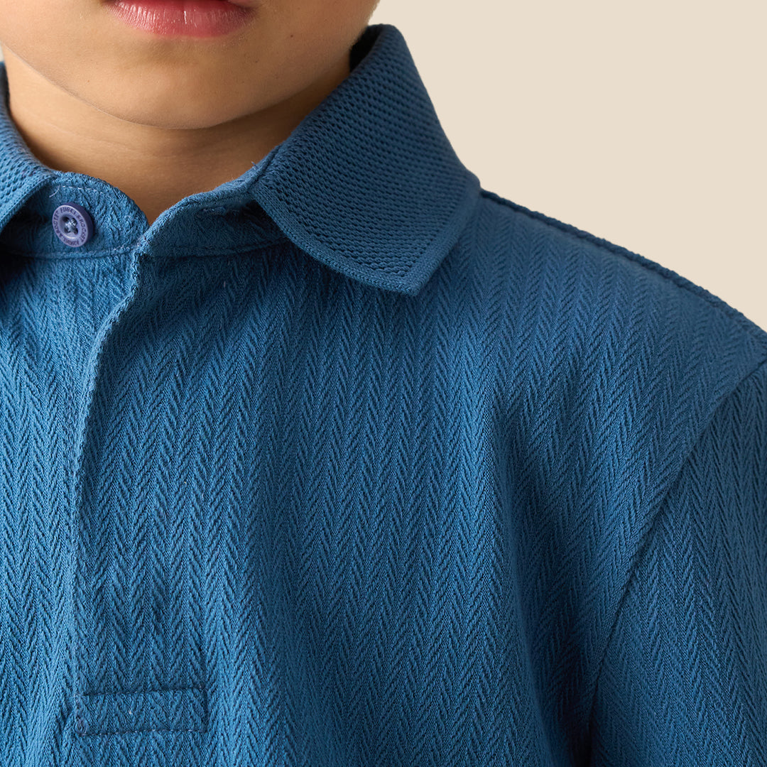 A young boy wearing a blue ribbed long-sleeve polo shirt and brown pants, standing against a solid beige background.