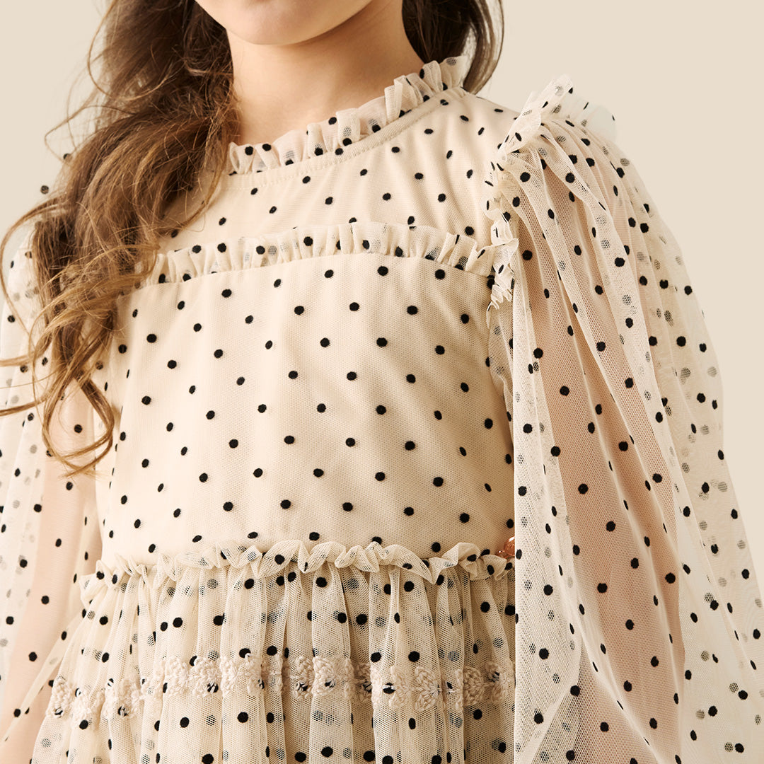 A young girl with her hair in a bun wears a cream-colored, long-sleeved tiered dress with black polka dots, standing against a solid beige background.