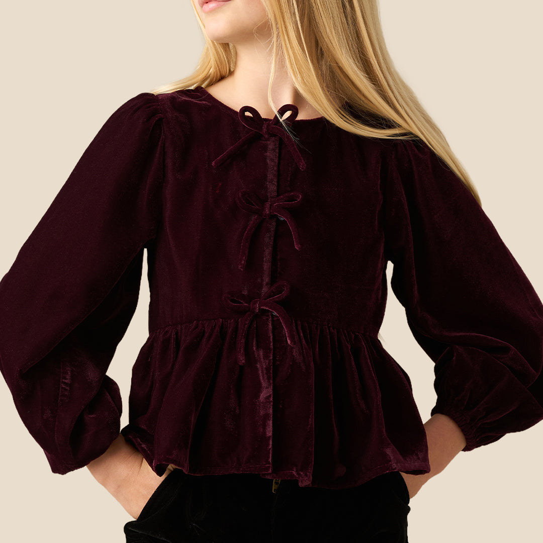 A young girl with long blonde hair wearing a deep burgundy velvet long-sleeve top with a tie-neck and peplum hem, paired with black pants, set against a solid light beige background.