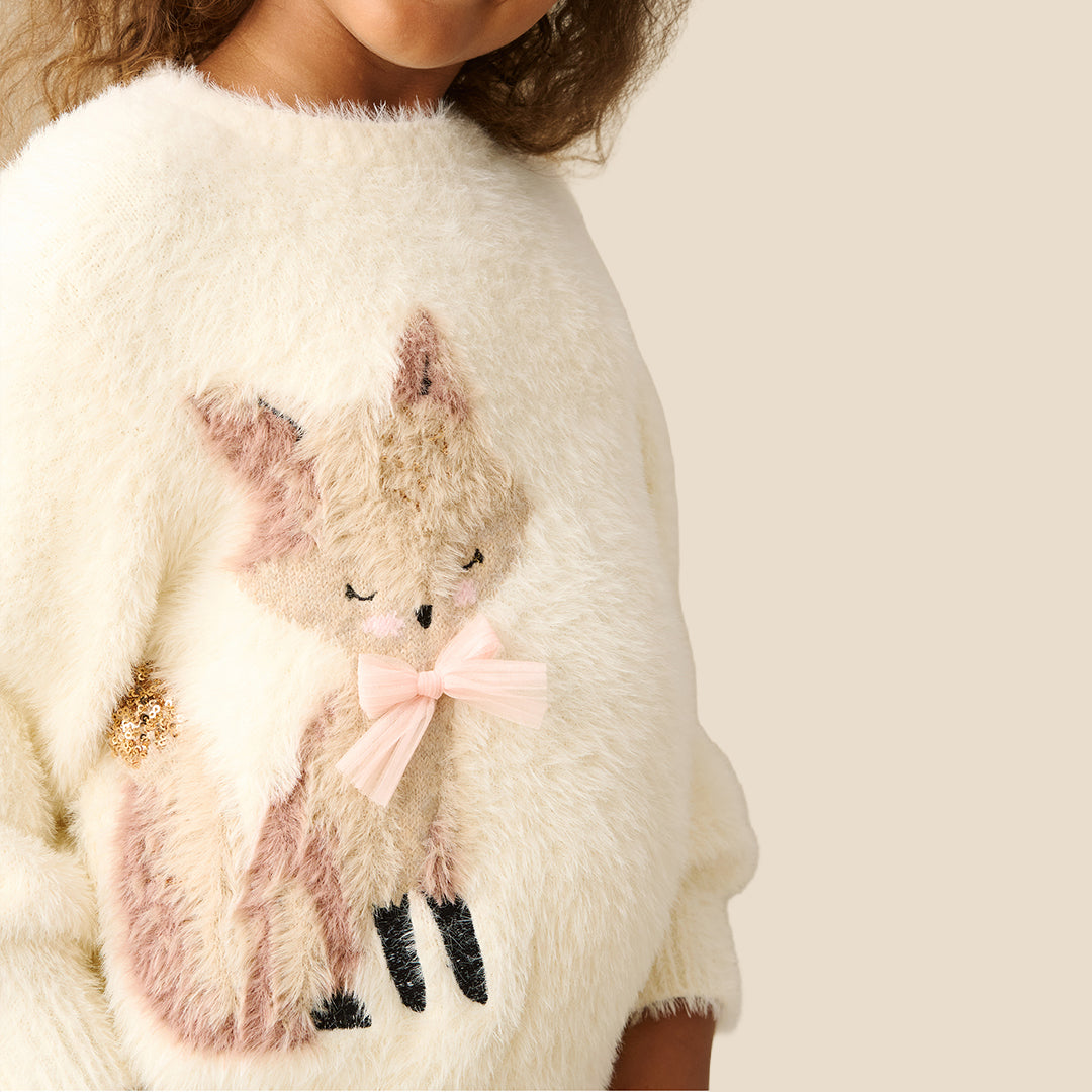 A young girl with curly brown hair wearing a fuzzy off-white knit sweater featuring a sequined fox applique on the front, paired with a gold sequined skirt, set against a solid light beige background.