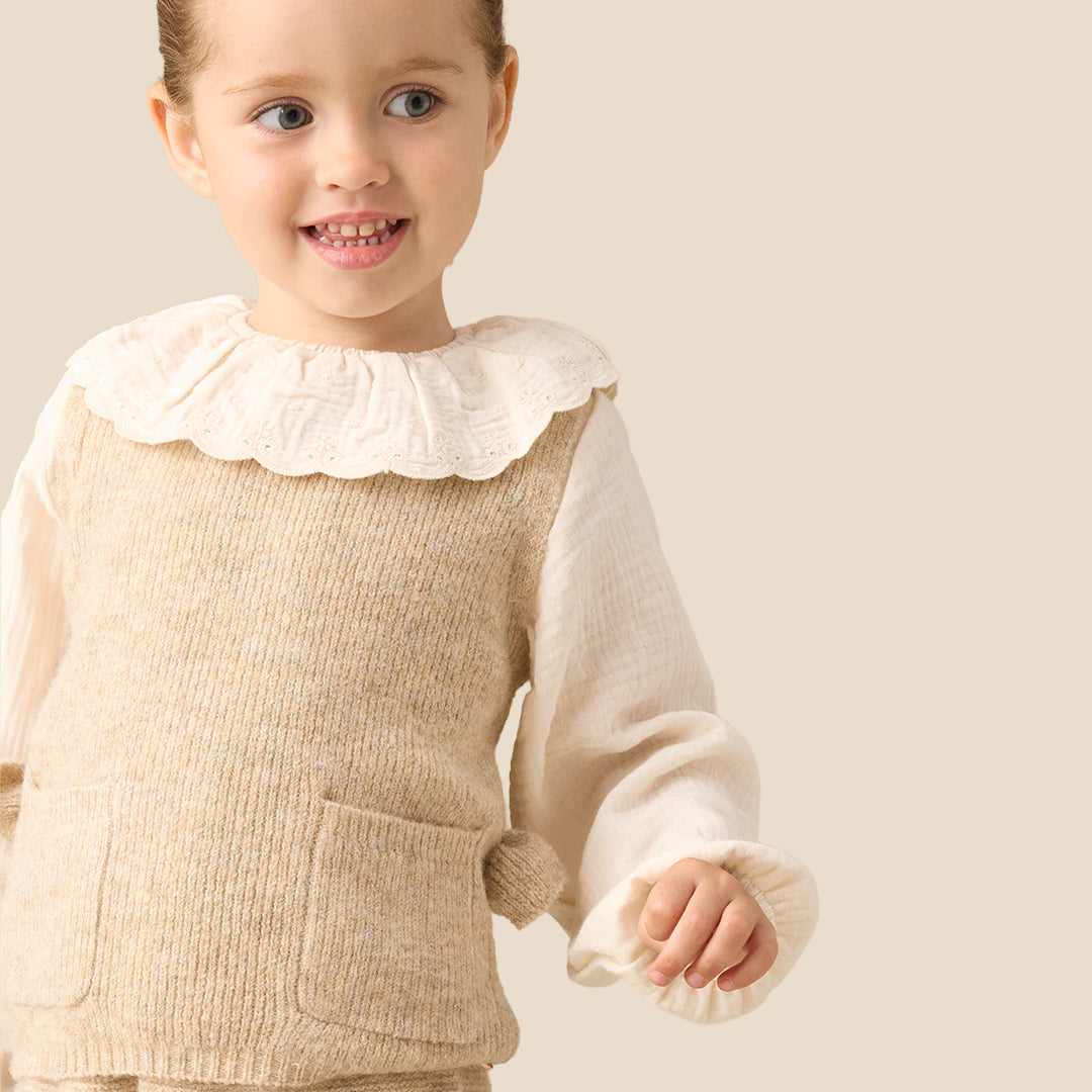 A young child modeling a cream and beige knit outfit, consisting of a long-sleeve top with a ruffled collar and matching knit vest and pants, set against a solid light beige background.