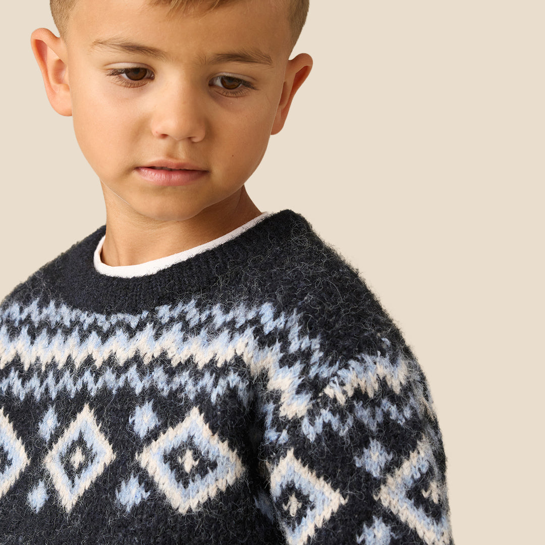 A young boy wearing a dark navy knit sweater with a white and light blue geometric Fair Isle pattern across the chest and shoulders, paired with beige pants, set against a solid light beige background.